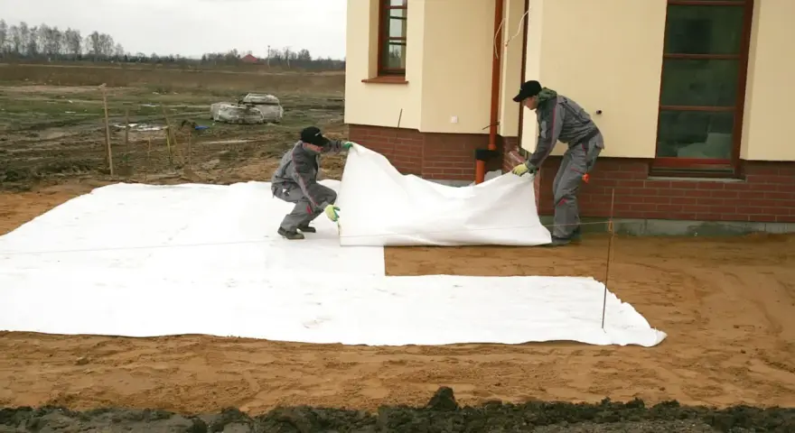 Dwóch robotników rozkłada białą geowłókninę, przygotowując tani taras na gruncie.