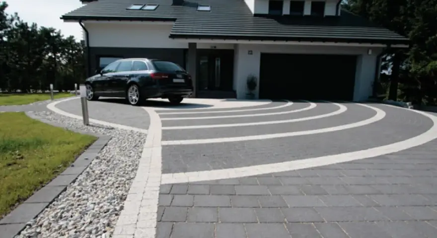 Nowoczesny dom z garażem, przed którym rozciąga się efektowny beton na podjazd, ozdobiony białymi pasami tworzącymi łuki.