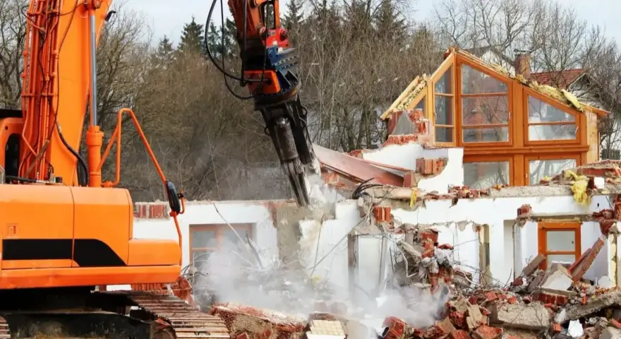 Pomarańczowa koparka niszczy dom, unosząc tumany kurzu i gruzu.