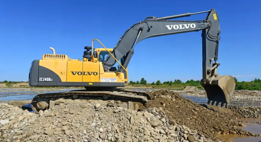 Żółta koparka Volvo EC240B LC pracuje na kamienistym brzegu rzeki. Zastanawiasz się, ile kosztuje godzina pracy koparki?