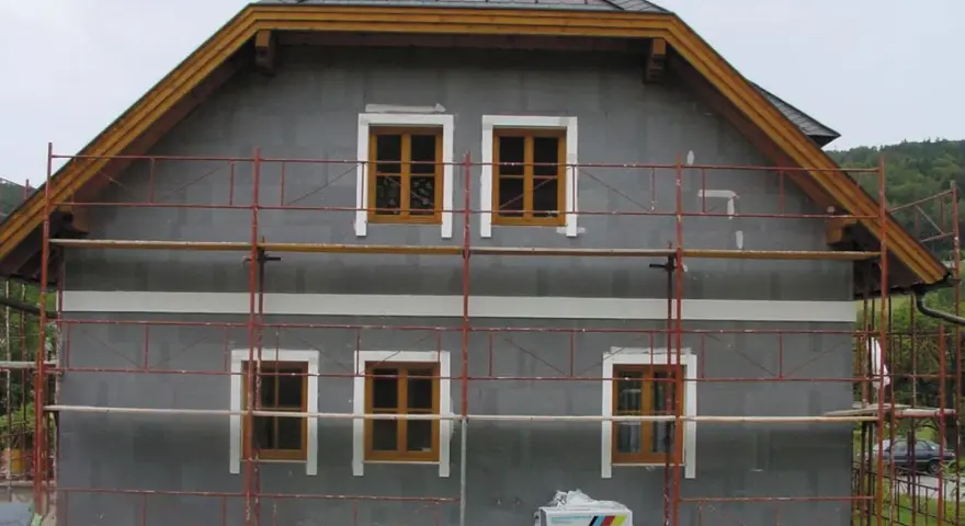 Dom w trakcie ocieplania. Szara elewacja, drewniane okna, rusztowanie. Materiały budowlane Austrotherm wskazują na dbałość o niski współczynnik przewodzenia ciepła.