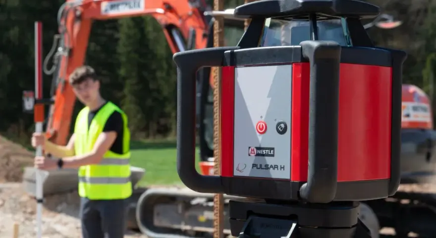 Niwelator laserowy Pulsar H na budowie. W tle pracownik w kamizelce odblaskowej i koparka.