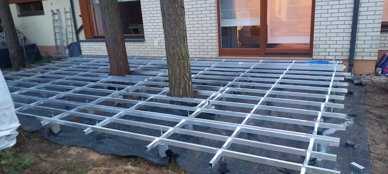 Konstrukcja tarasu z deski kompozytowej bez legarów, z drzewami przechodzącymi przez ruszt.