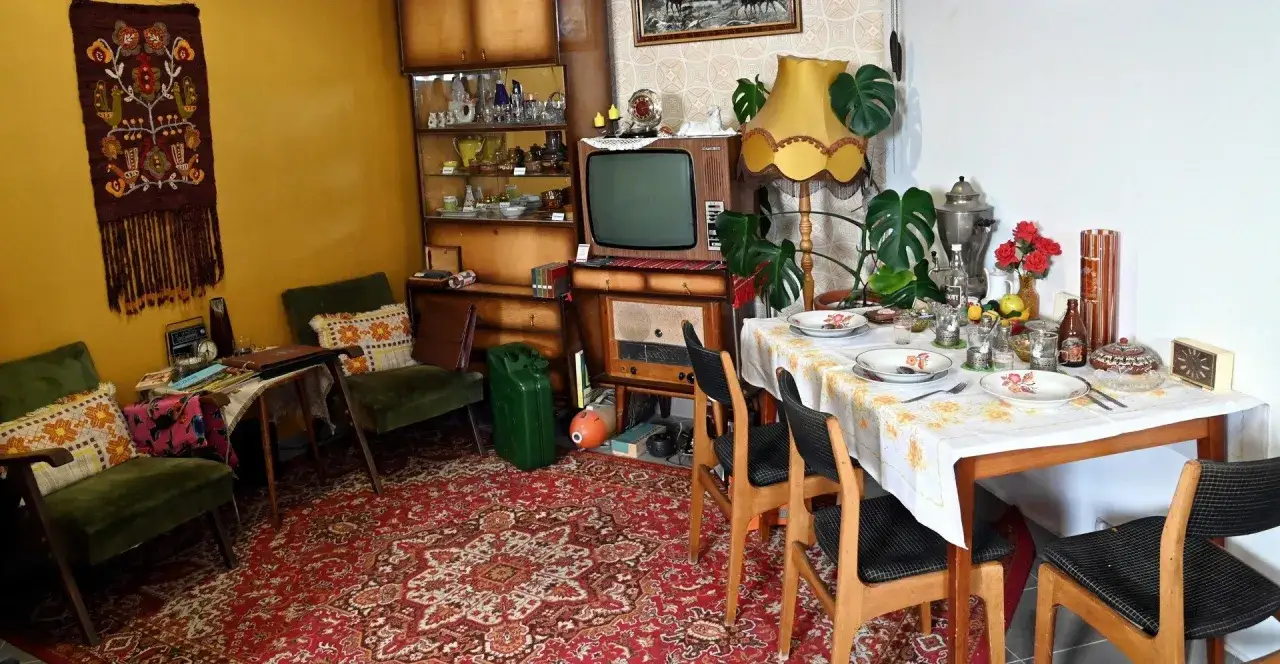 Salon w stylu prl: zielone fotele, dywan, meblościanka z bibelotami, telewizor, radio i stół nakryty do obiadu.