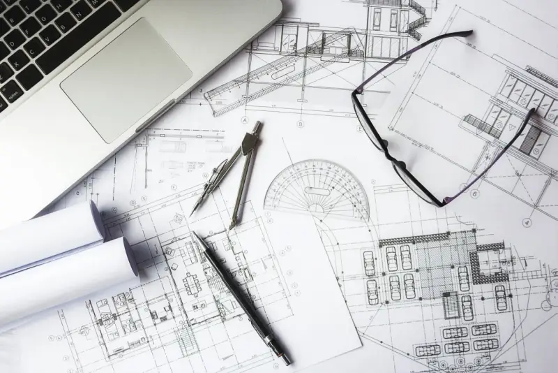 Na stole leżą plany architektoniczne, laptop, okulary i przybory kreślarskie. To miejsce pracy nad kosztorysem inwestorskim.
